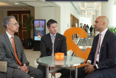 Three business professionals seated around a small round table in a modern conference venue, engaged in discussion. They are wearing formal suits with Janus Henderson branding, with an orange sculptural logo and digital display in the background, suggesting a professional interview or investor discussion setting.