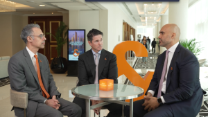 Three business professionals seated around a small round table in a modern conference venue, engaged in discussion. They are wearing formal suits with Janus Henderson branding, with an orange sculptural logo and digital display in the background, suggesting a professional interview or investor discussion setting.