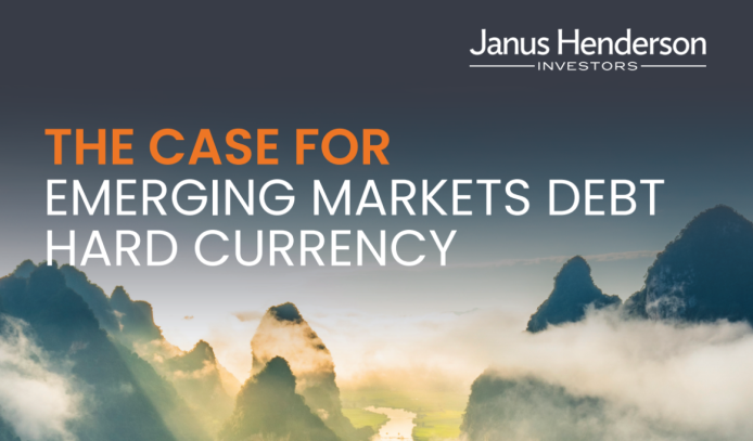Cover image for a Janus Henderson Investors publication titled “The Case for Emerging Markets Debt Hard Currency.” The title is displayed prominently in large uppercase text, with “THE CASE FOR” in orange and “EMERGING MARKETS DEBT HARD CURRENCY” in white. The Janus Henderson Investors logo appears in the top-right corner. The background shows a dramatic mountain landscape with steep, mist-covered peaks rising above clouds, illuminated by soft sunrise or sunset light. The sky transitions from dark blue at the top to pale light near the horizon, creating a calm, atmospheric scene.