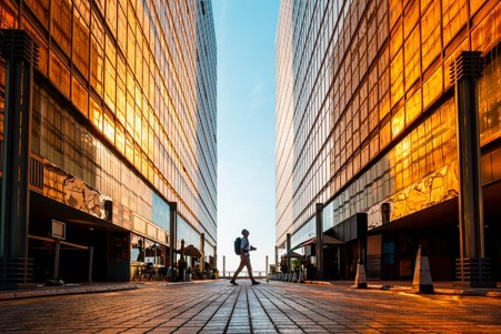 A lone pedestrian walks across a paved plaza between two tall glass office buildings. The buildings reflect warm golden sunlight, creating a symmetrical corridor effect, while the low-angle perspective emphasizes the height of the towers and the open sky visible between them.