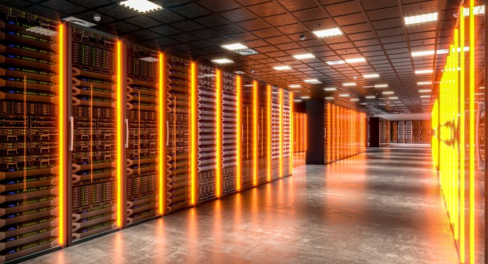 A wide corridor inside a modern data centre lined with tall server racks on both sides. The servers emit vertical lines of orange and amber light, reflecting off a polished concrete floor. Overhead ceiling panels extend into the distance, creating strong linear perspective that emphasises scale, infrastructure, and advanced digital technology.