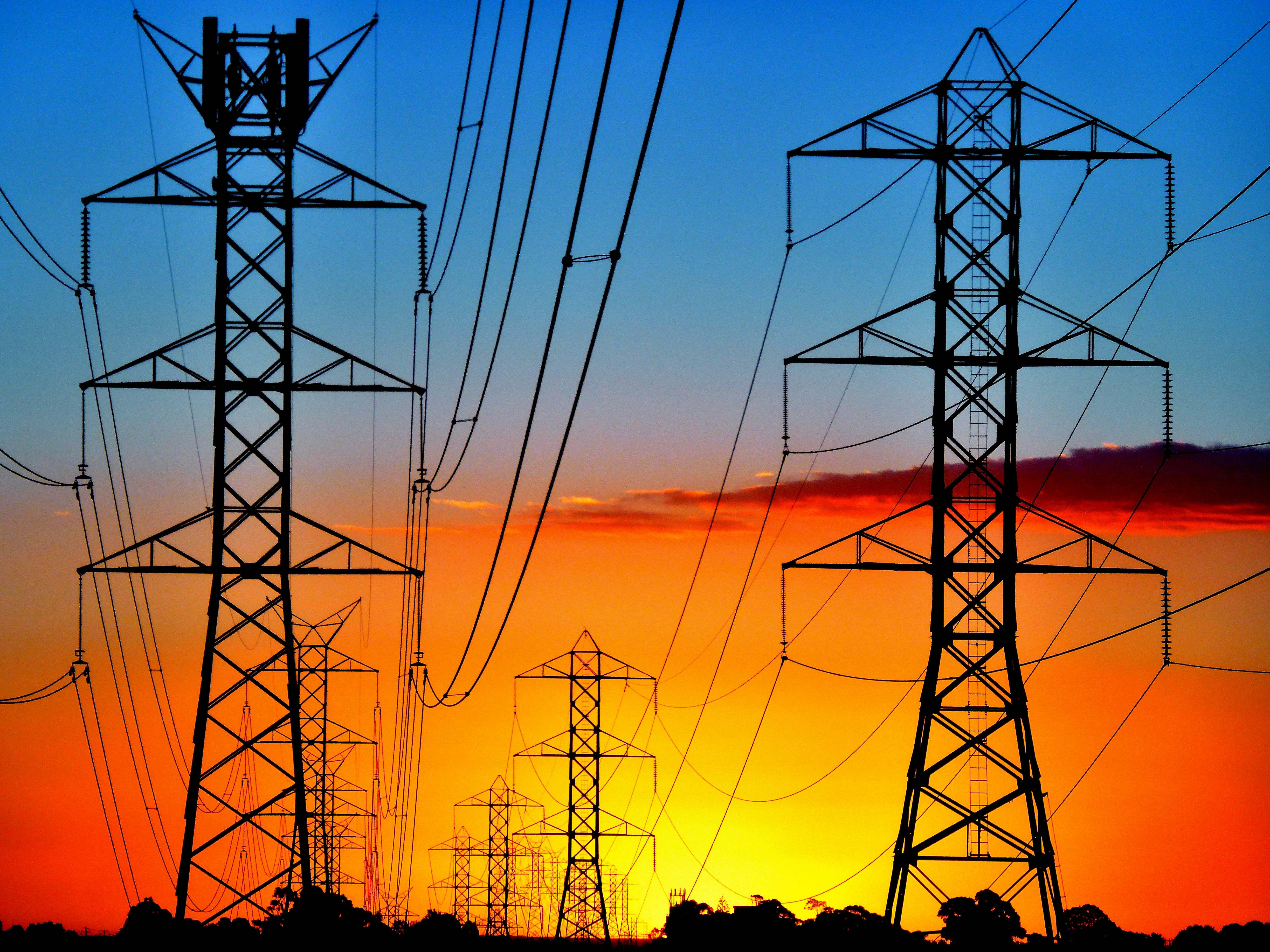 Electricity transmission pylons and high-voltage power lines silhouetted against a vivid orange and blue sunset sky, with multiple towers receding into the distance.