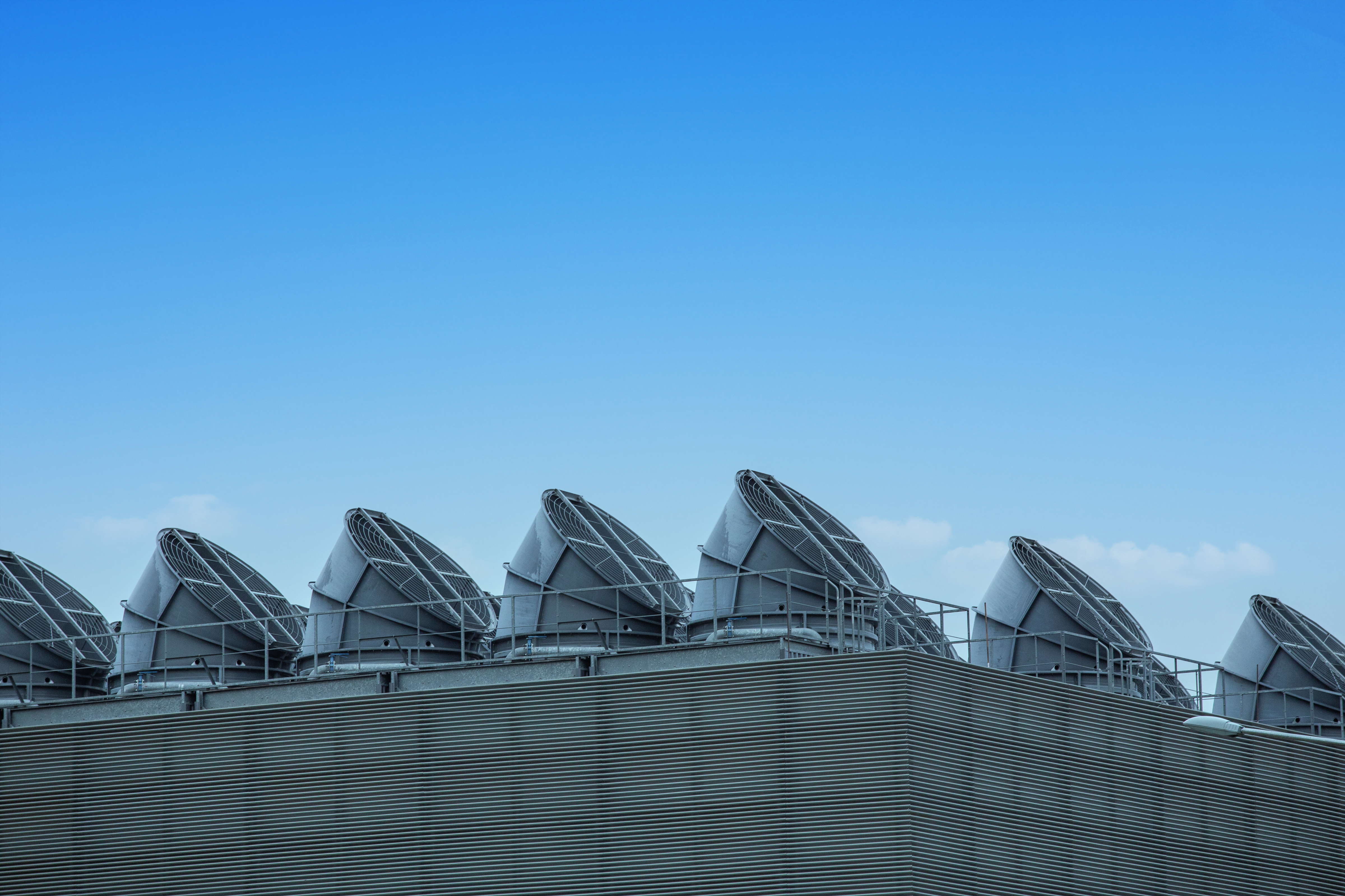 Row of large rooftop cooling and ventilation units on a modern industrial building, with metal louvred cladding and a clear blue sky in the background. The image highlights the cooling infrastructure used to regulate temperatures in data centres and other high-performance facilities.