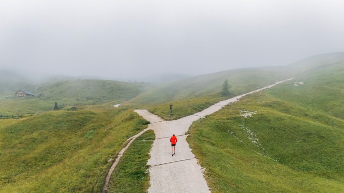 A lone runner wearing a bright red jacket stands at the centre of a forked gravel path in rolling green hills. The paths diverge left and right into mist, with visibility fading into a pale, overcast sky. The surrounding landscape is open and softly textured, suggesting choice, uncertainty, and decision making at a crossroads.