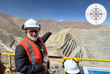 A person in safety gear points across a large open‑pit mine carved into a mountainous, arid landscape, with terraced rock walls stretching into the Andes under a clear blue sky; a compass graphic appears in the top corner indicating direction.