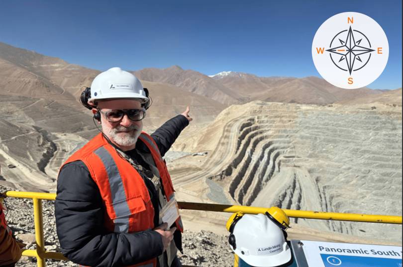 A person in safety gear points across a large open‑pit mine carved into a mountainous, arid landscape, with terraced rock walls stretching into the Andes under a clear blue sky; a compass graphic appears in the top corner indicating direction.