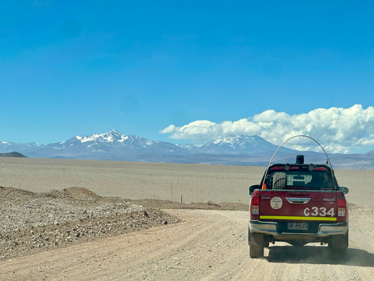 Red off‑road vehicle driving along a dusty desert road with snow‑capped Andes mountains under a clear blue sky.