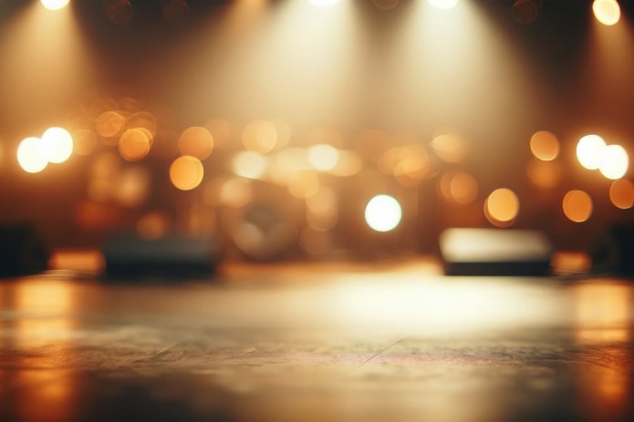 An empty stage viewed from floor level, softly out of focus, with warm amber and golden lights glowing in the background. Circular bokeh highlights suggest stage spotlights and lighting rigs, while microphone stands and instruments are faintly visible. The image conveys anticipation, readiness, and performance without an audience present.