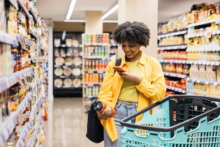 Eine Person, die in dem Gang eines Supermarkts einkauft, ein verpacktes Lebensmittel und eine wiederverwendbare Wasserflaschen in der Hand hält, während sie neben einem Einkaufskorb steht, an beiden Seiten haben wir Ablagen mit Lebensmitteln.